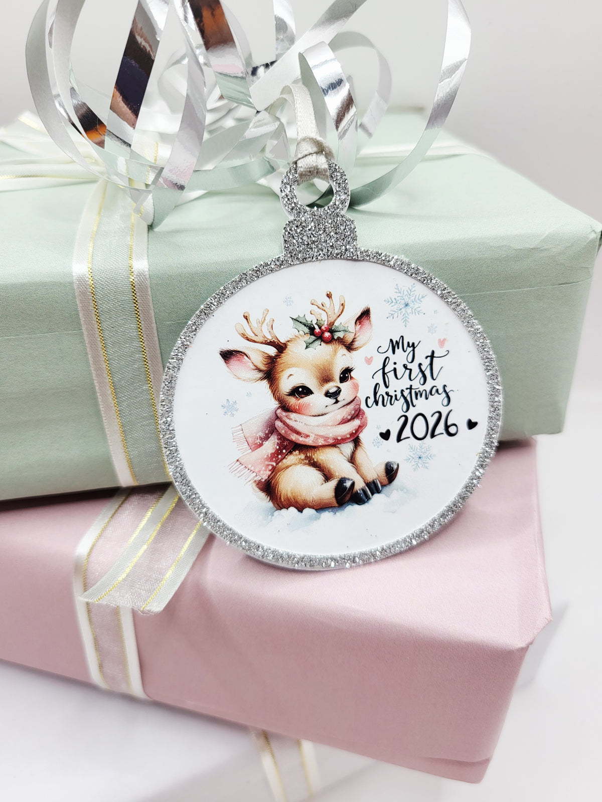 A round Christmas gift tag in the shape of a bauble, featuring a cute baby reindeer wearing a pink scarf, with the text 'My first Christmas 2026'. The gift tag has a glittery silver border and a silver satin ribbon to attach to your gift.