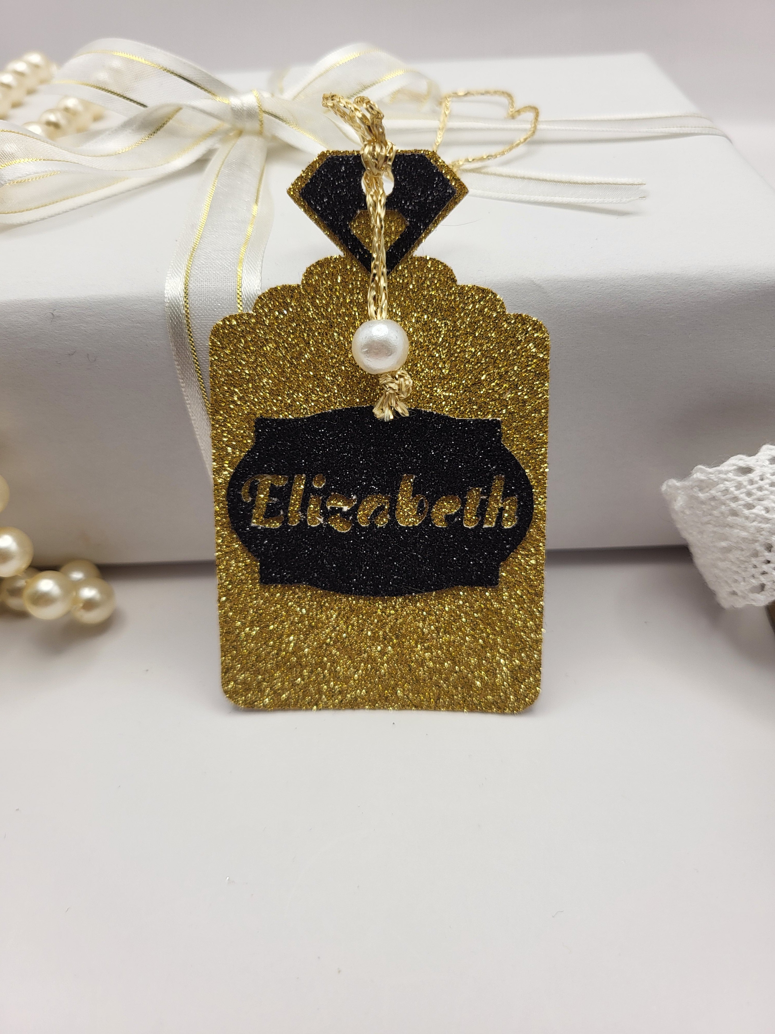 Gold glitter card Perfume bottle shape gift tag has a black tag shape stuck on the front with the chance to personalise it with the name of your choice. The bottle top is also black glitter card with a metallic gold cord attached