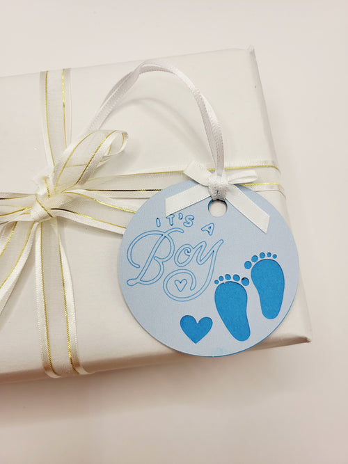 It's a boy gift tag is pale blue with two little feet and a small love heart cut out. It is stuck to a darker blue back card and this shows through the cut out feet and heart. The words It's  a boy are written on in a blueberry colour. The tag has a white bow and a white satin ribbon to attach the tag to a gift.
