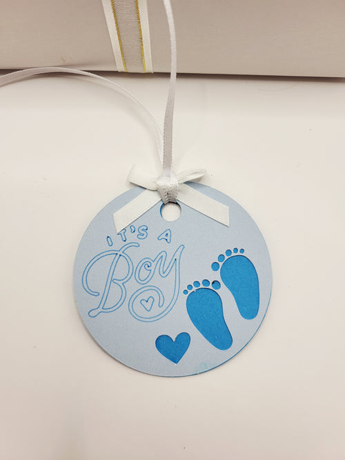 It's a boy gift tag is pale blue with two little feet and a small love heart cut out. It is stuck to a darker blue back card and this shows through the cut out feet and heart. The words It's  a boy are written on in a blueberry colour. The tag has a white bow and a white satin ribbon to attach the tag to a gift.