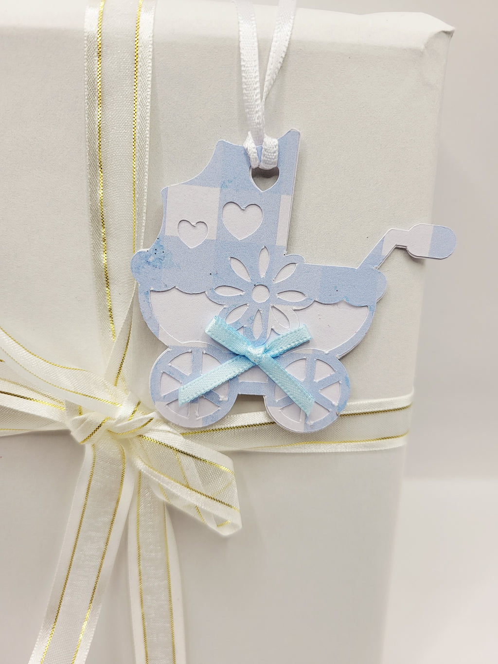 Pram shaped gift tag in blue gingham  on a white background. Pram has a blue bow and a white satin ribbon to attach the tag