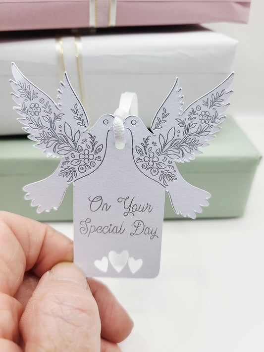 A white card gift tag with the words On Your Special Day foiled in silver. There are tow doves one each side of the tag near the top. The pattern on the wings are also foiled. There are 3 love hearts cut out at the bottom of the tag and it is blank on the reverse 