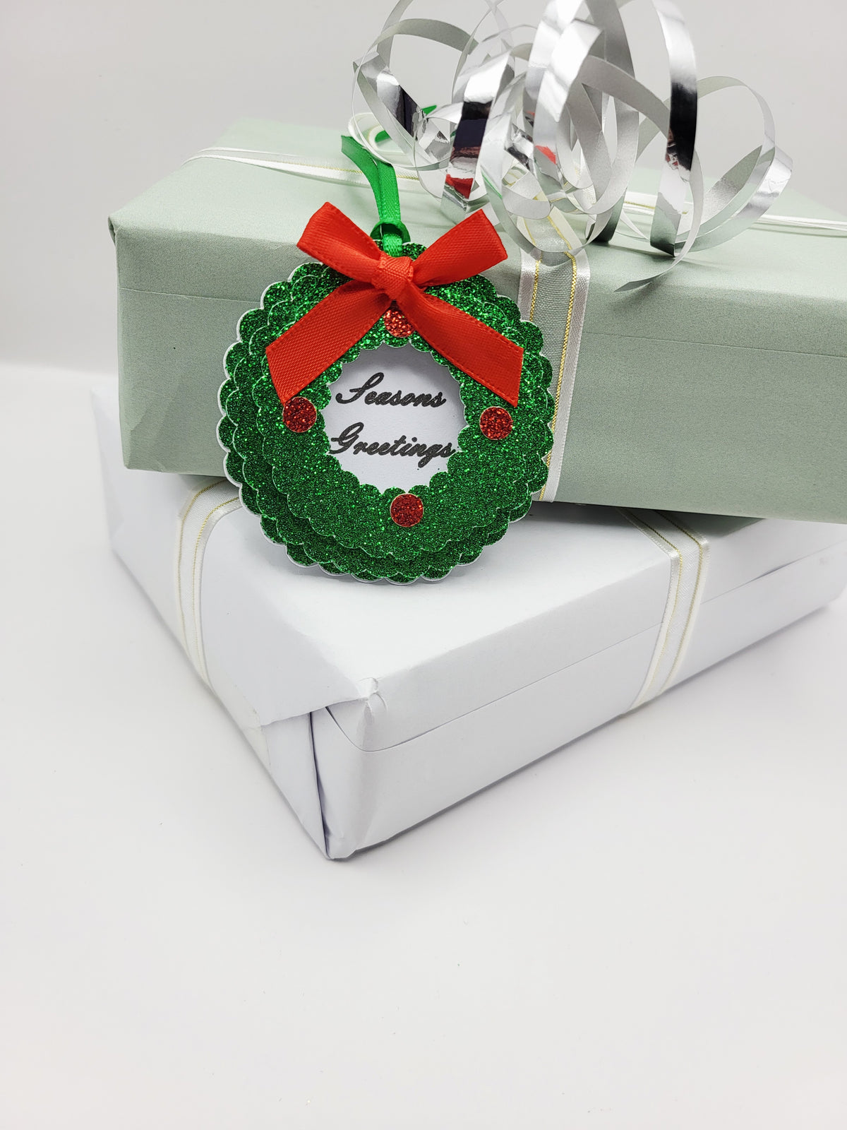 A green glitter Christmas wreath gift tag with a red ribbon bow and 'Seasons Greetings' text, in the centre on a white card background