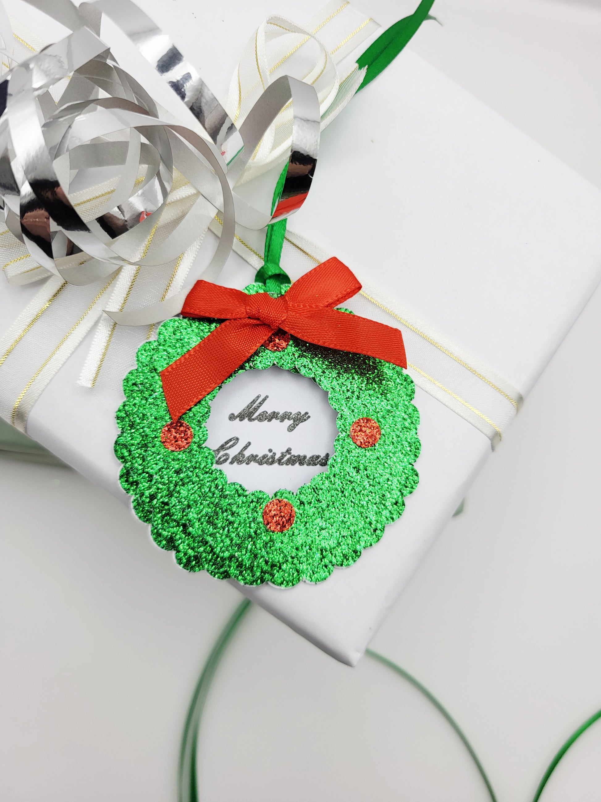 A green glitter Christmas wreath gift tag with a red ribbon bow and 'Merry Christmas' text, in the centre on a white card background