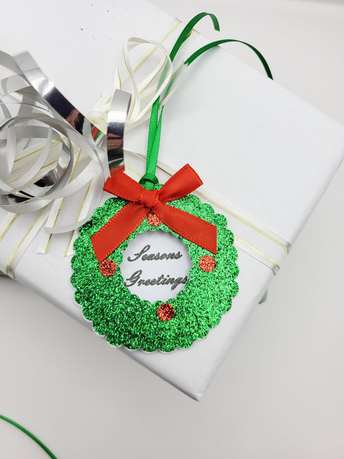 A green glitter Christmas wreath gift tag with a red ribbon bow and 'Seasons Greetings' text, in the centre on a white card background