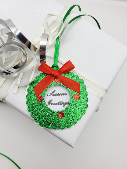 A green glitter Christmas wreath gift tag with a red ribbon bow and 'Seasons Greetings' text, in the centre on a white card background