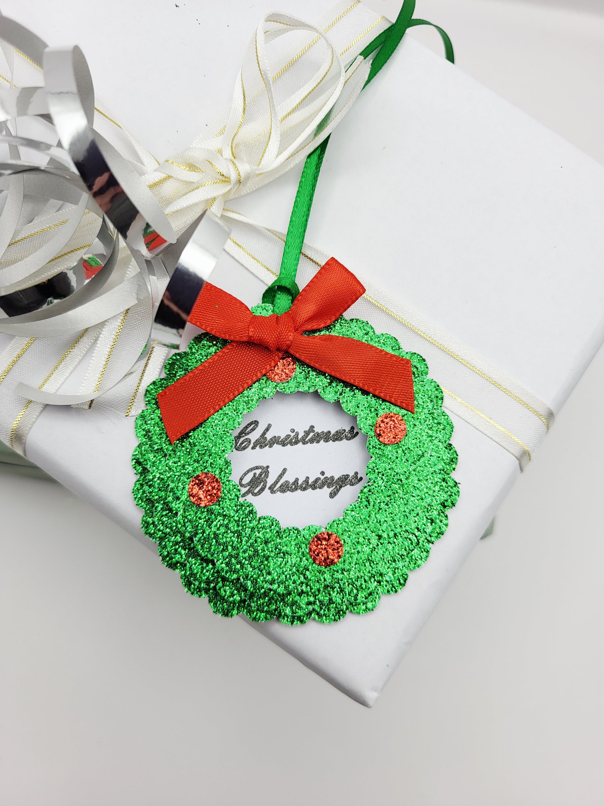 A green glitter Christmas wreath gift tag with a red ribbon bow and 'Christmas Blessings' text, in the centre on a white card background