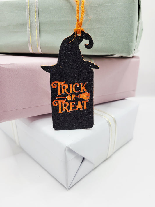 A black glitter witch hat shaped gift tag with orange 'Trick or Treat' lettering. It has a orange cord to attach to your gift