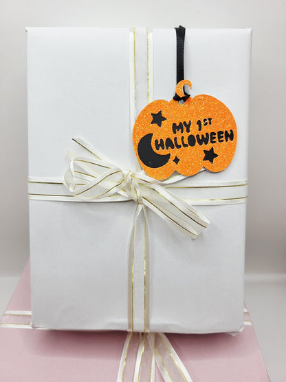 A  pumpkin-shaped Halloween Gift tag  with black lettering that reads 'MY 1ST HALLOWEEN' and black satin ribbon, to attach to your gift.