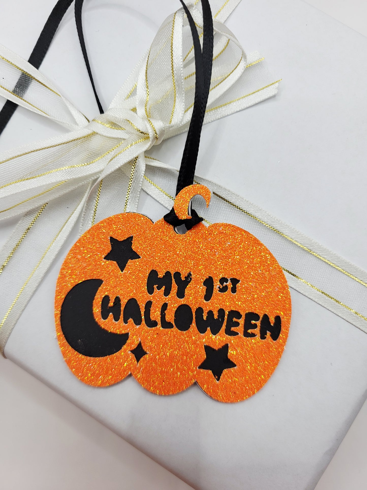 An orange glitter pumpkin-shaped gift tag with black lettering 'MY 1ST HALLOWEEN' and black star and moon graphics,  a black satin ribbon is attached to add to gift