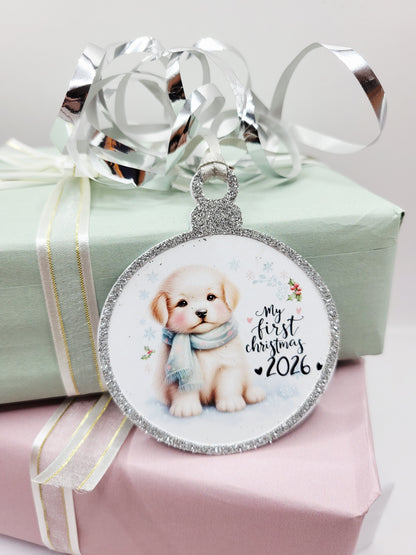 A round Christmas gift tag in the shape of a bauble, featuring a cute cream-colored puppy wearing a blue scarf, with the text 'My first Christmas 2026'. The gift tag has a glittery silver border and a silver satin ribbon to attach to your gift.

