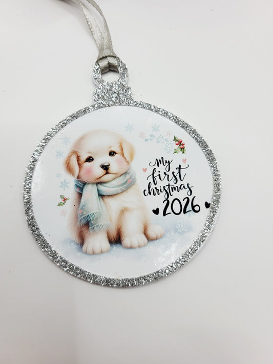 A round Christmas gift tag in the shape of a bauble, featuring a cute puppy illustration and the text 'my first christmas 2026' on a glittery silver border, with  a silver ribbon to attach to gift