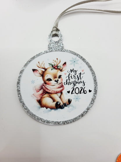 A round Christmas gift tag in the shape of a bauble, featuring a cute baby reindeer illustration and the text 'my first christmas 2026' on a glittery silver border, with  a silver ribbon to attach to gift