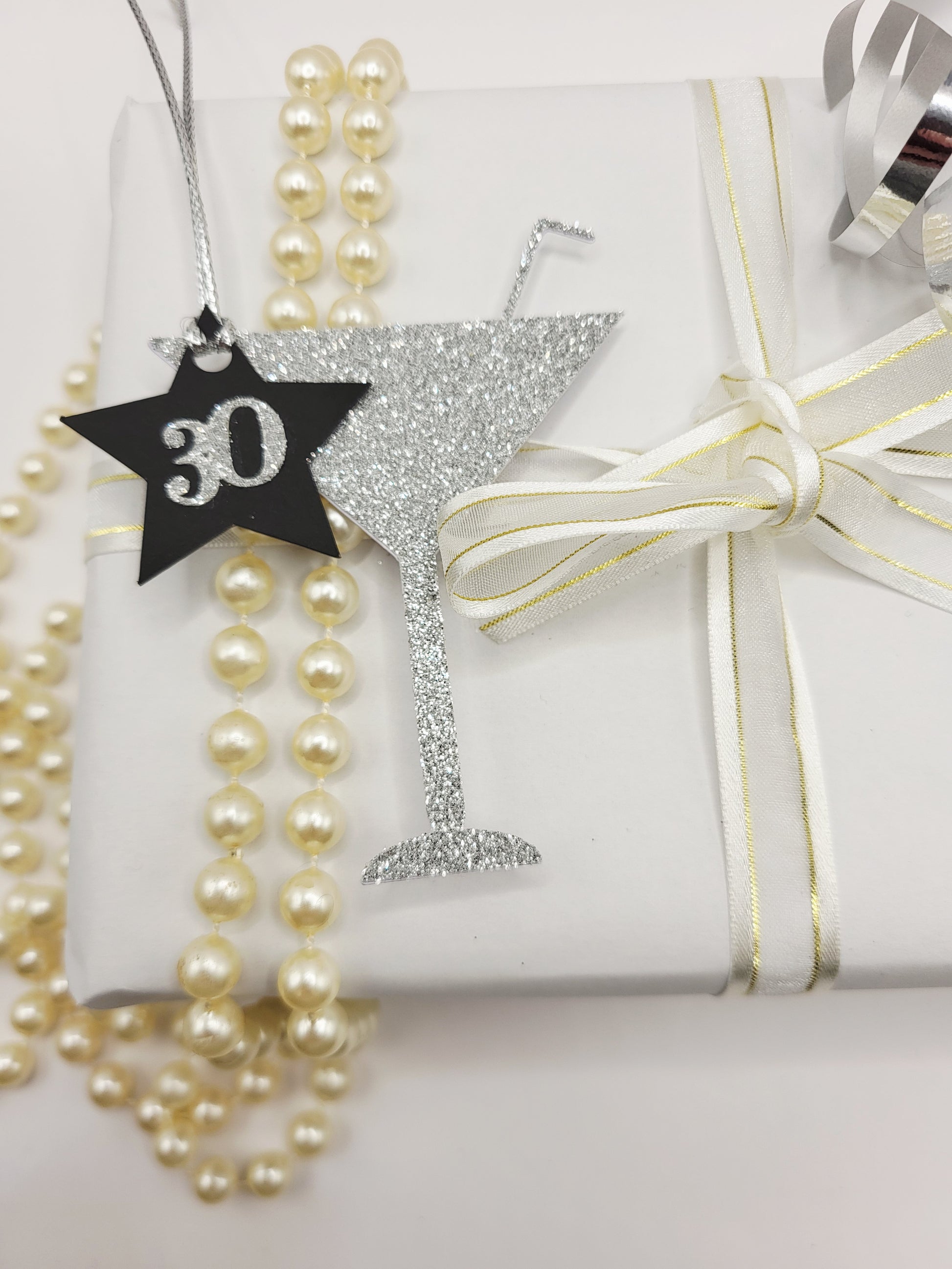 Milestone Cocktail glass gift tag is made from silver glitter with a straw sticking from the glass. The birthday age is on a black card star with the number glued onto the star. There is a silver metallic cord to attach the tag to the gift.