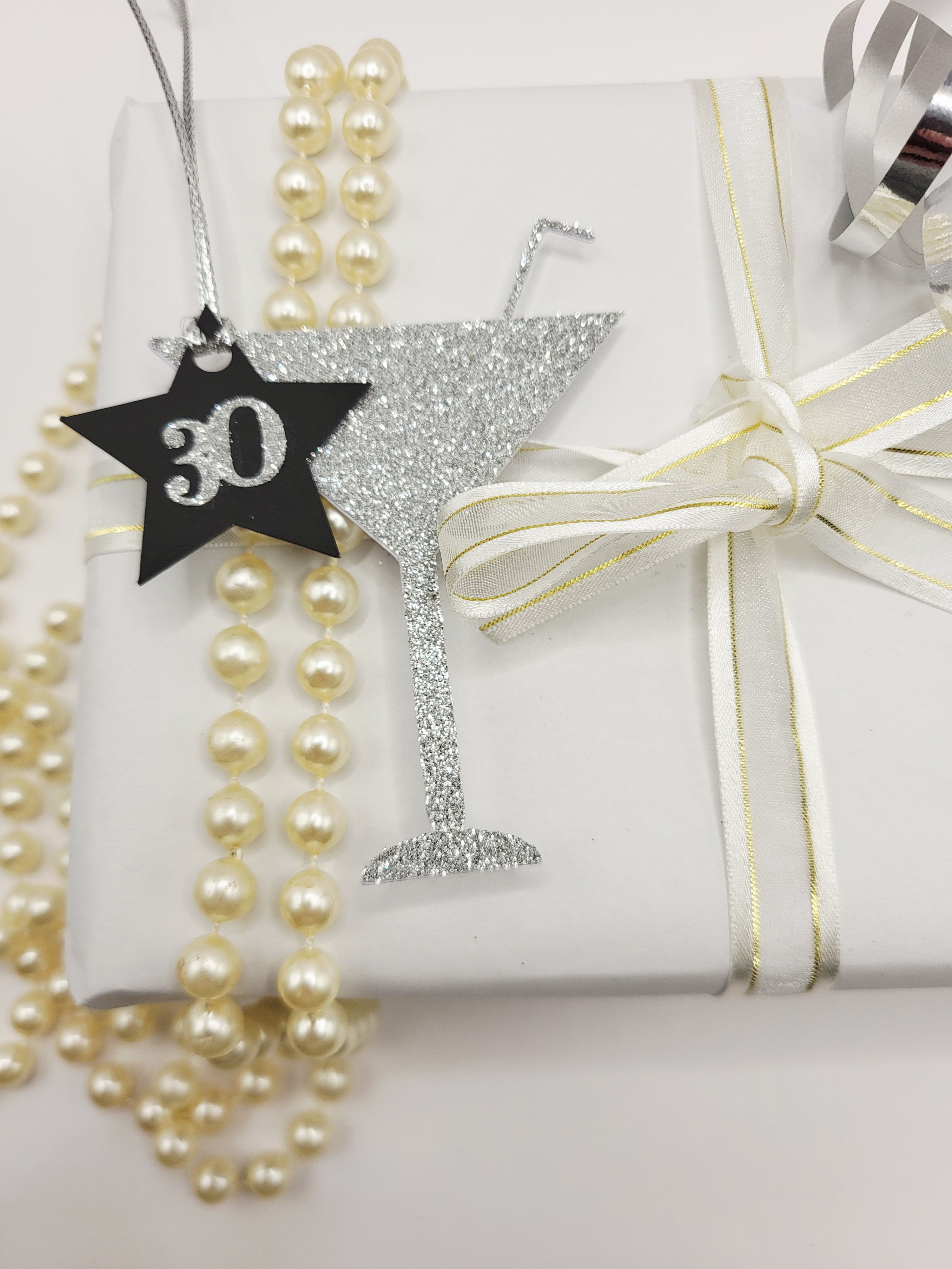 Milestone Cocktail glass gift tag is made from silver glitter with a straw sticking from the glass. The birthday age is on a black card star with the number glued onto the star. There is a silver metallic cord to attach the tag to the gift.