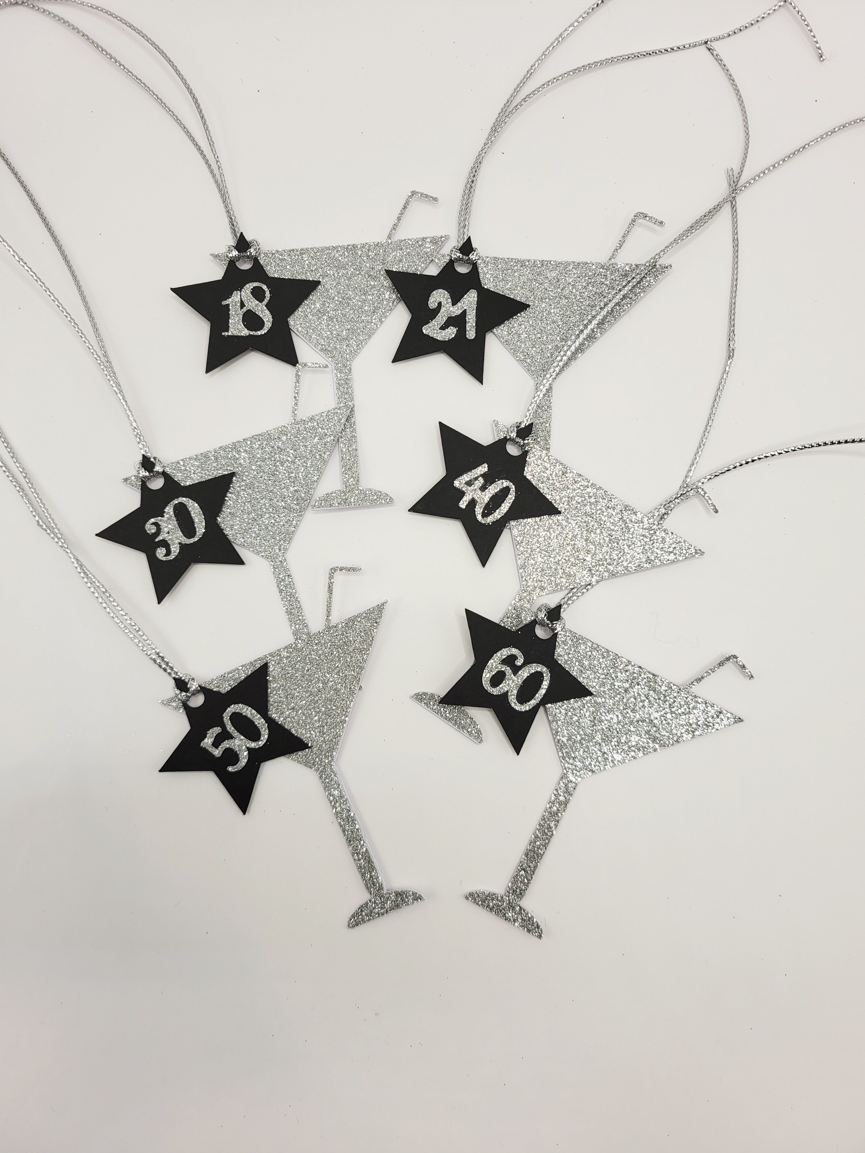6 lots of silver glitter milestone age cocktail shaped gift tags. The glass shapes have a straw sticking from the top.
There is a black card star with the milestone ages in silver glitter stuck on the star and hung from the glass. The tags have a silver metallic cord attached so you can attach your gift easily