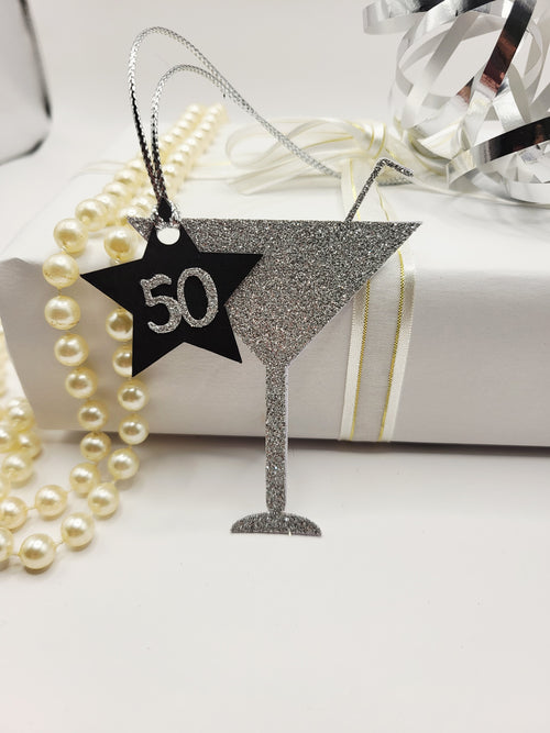 Milestone Cocktail glass gift tag is made from silver glitter with a straw sticking from the glass. The birthday age is on a black card star with the number glued onto the star. There is a silver metallic cord to attach the tag to the gift.