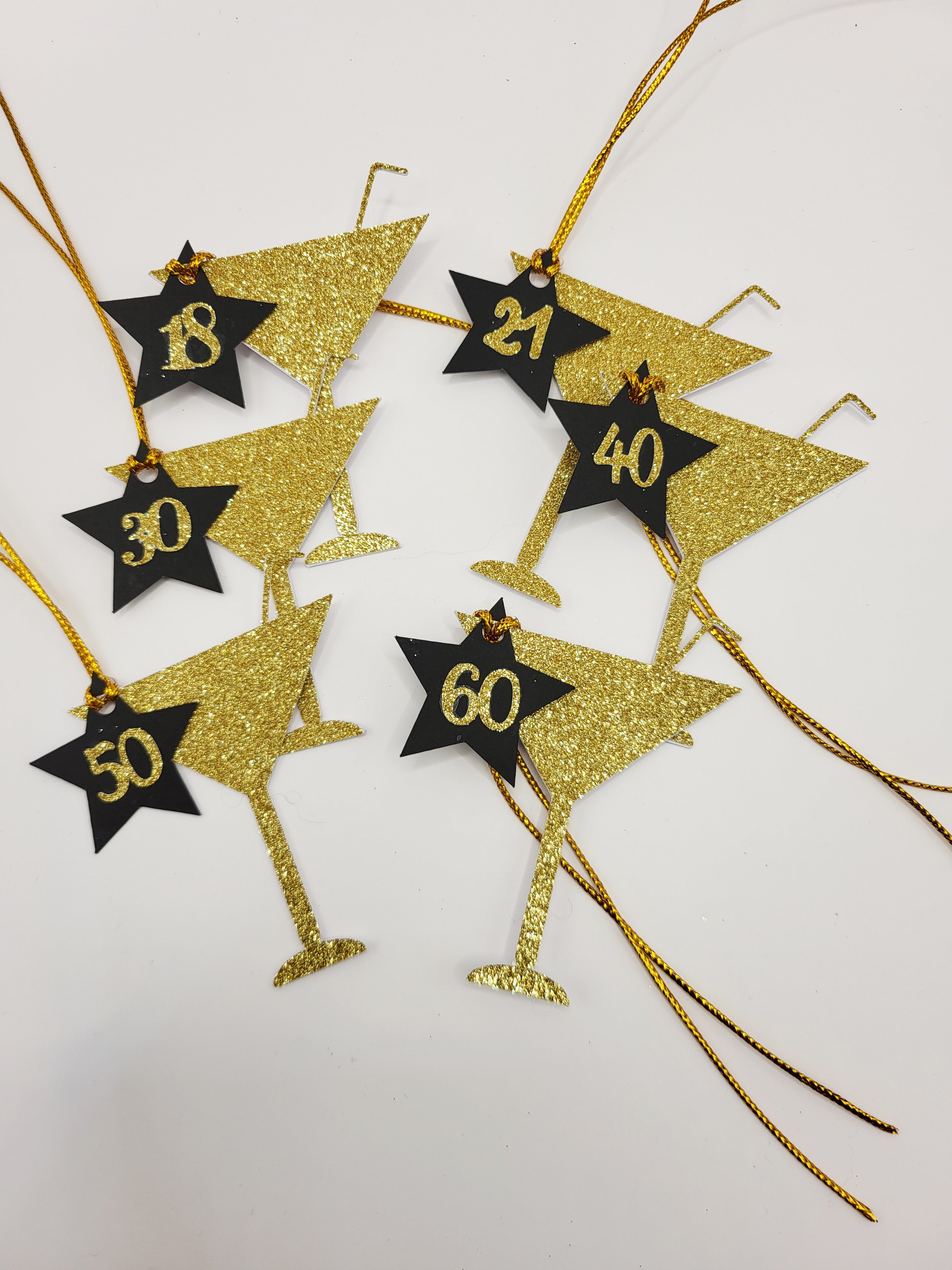 6 lots of Gold glitter milestone age cocktail shaped gift tags. The glass shapes have a straw sticking from the top.
There is a black card star with the milestone ages in gold glitter stuck on the star and hung from the glass. The tags have a gold metallic cord attached so you can attach your gift easily