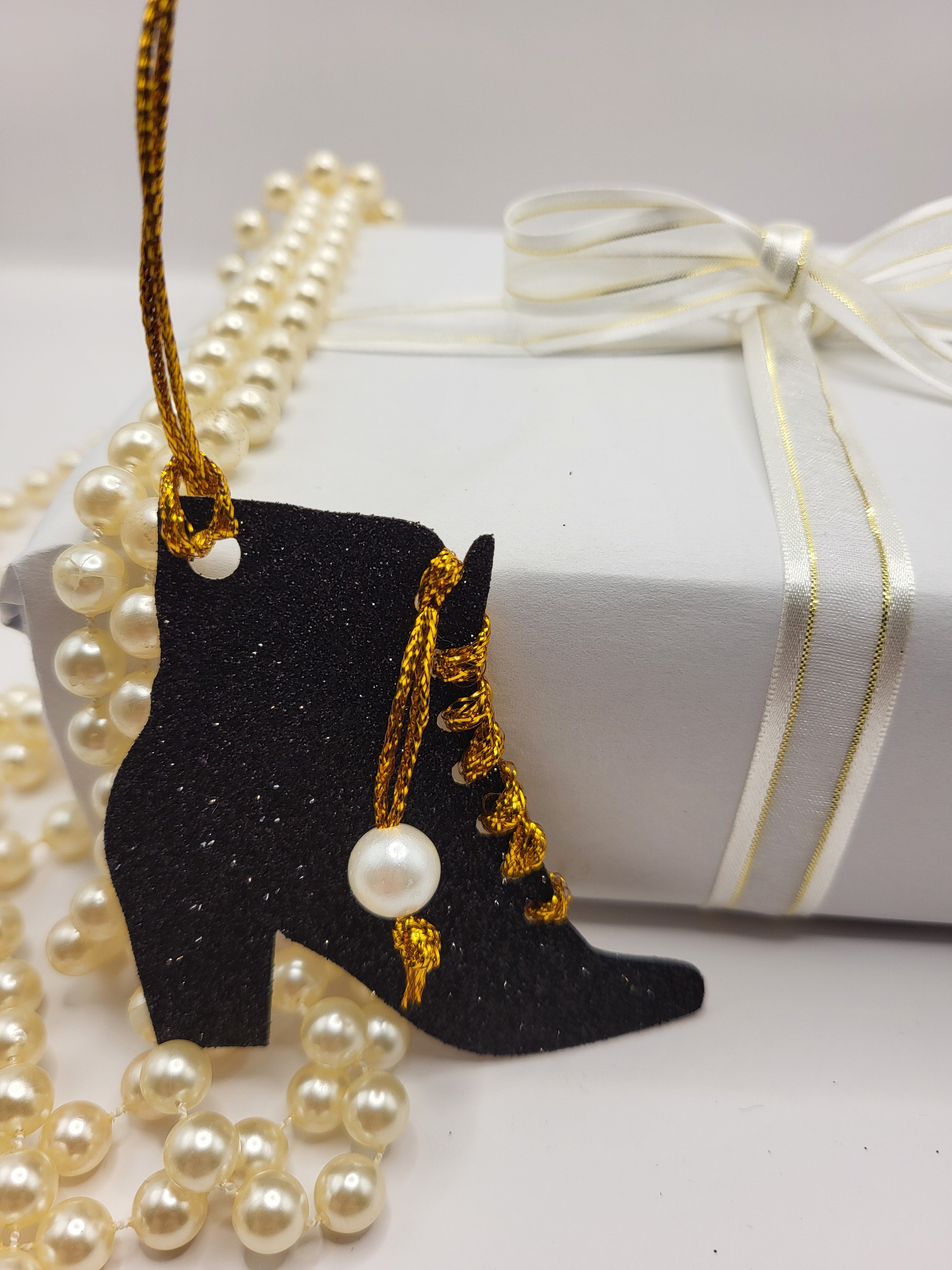 This is a black glitter ankle boot shaped gift tag with gold metallic cord threaded through the eyelets with a white pearl hanging from the thread.
