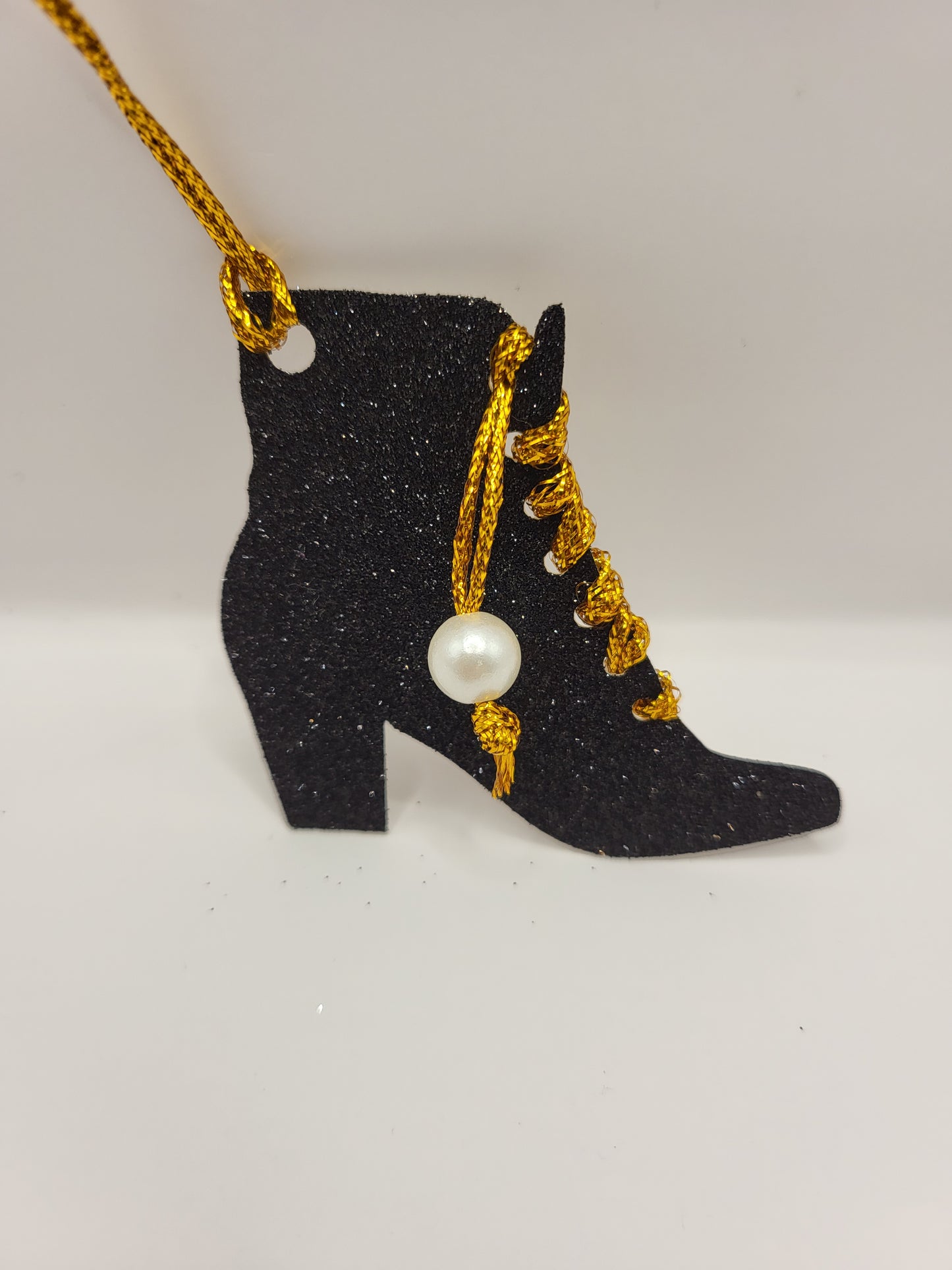 This is a black glitter ankle boot shaped gift tag with gold metallic cord threaded through the eyelets with a white pearl hanging from the thread.