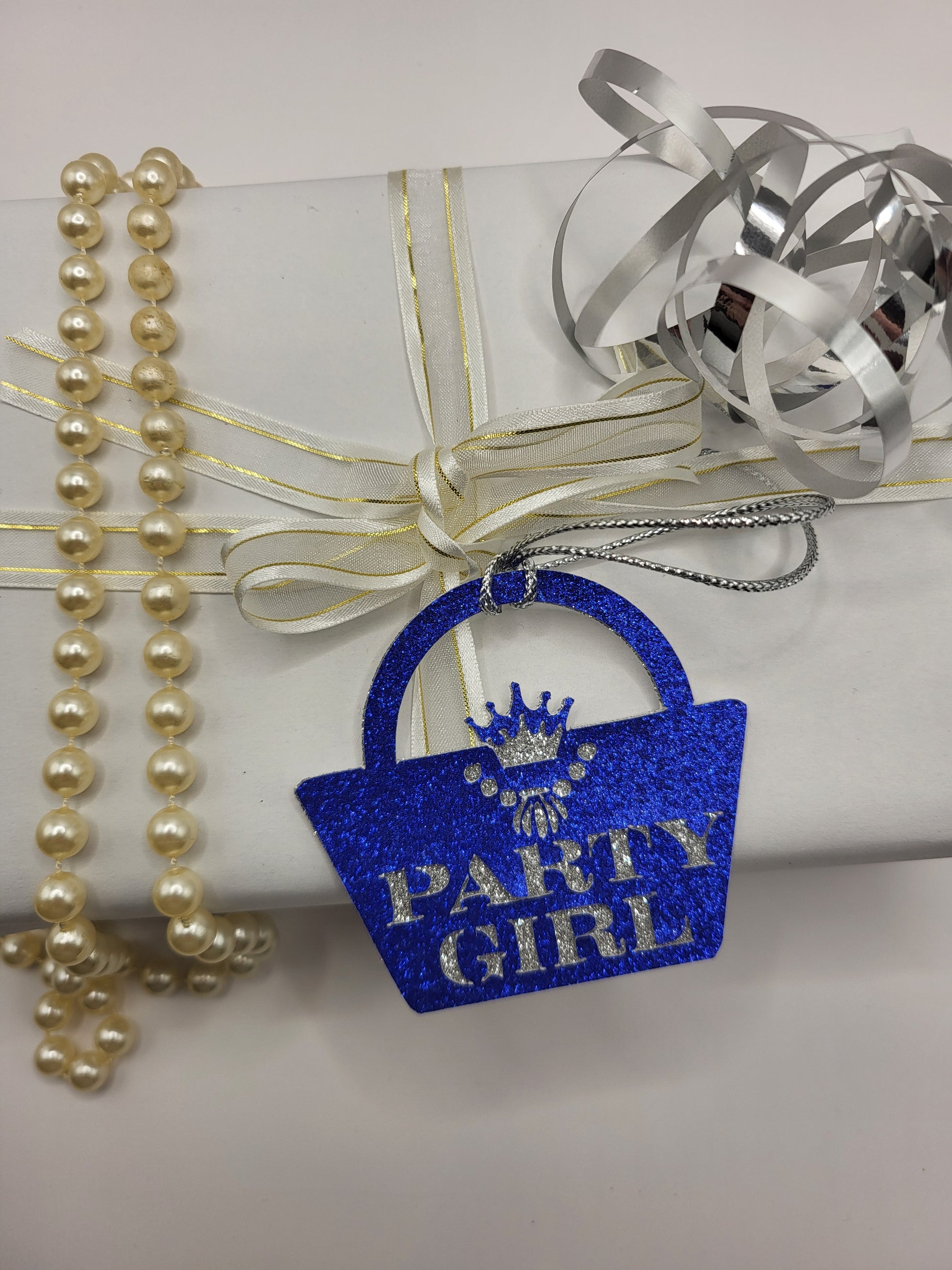 Blue glitter card handbag shaped gift tag stuck to a silver glitter background. The words party girl are cut out of the blue glitter card and shows the silver glitter underneath. The handbag clasp is the shape of the crown and there is a necklace cut out under the crown