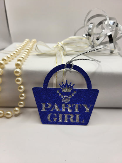 Blue glitter card handbag shaped gift tag stuck to a silver glitter background. The words party girl are cut out of the blue glitter card and shows the silver glitter underneath. The handbag clasp is the shape of the crown and there is a necklace cut out under the crown