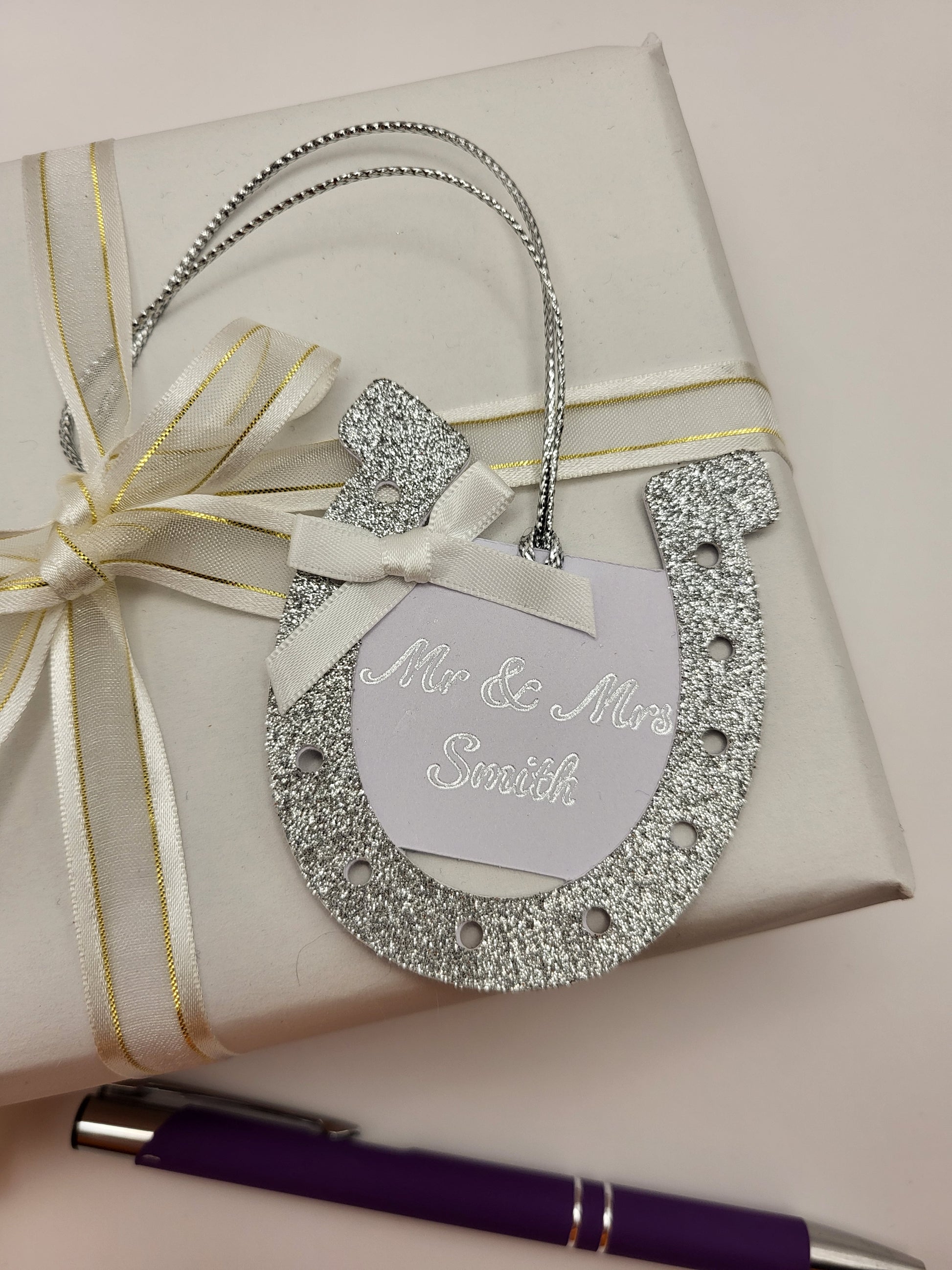 silver glitter card horseshoe shape gift tag. There is a white card centre with the words Mr &  Mrs Smith. This can be personalised into any surname you require. There is a white bow at the top left had side of the horseshoe. There is a silver metallic cord attached so you can attach it to your gift