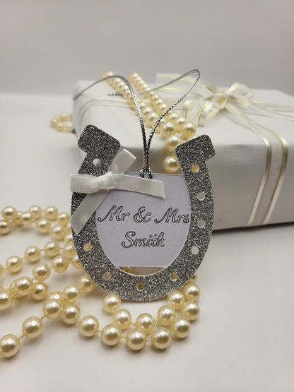 silver glitter card horseshoe shape gift tag. There is a white card centre with the words Mr &  Mrs Smith. This can be personalised into any surname you require. There is a white bow at the top left had side of the horseshoe. There is a silver metallic cord attached so you can attach it to your gift