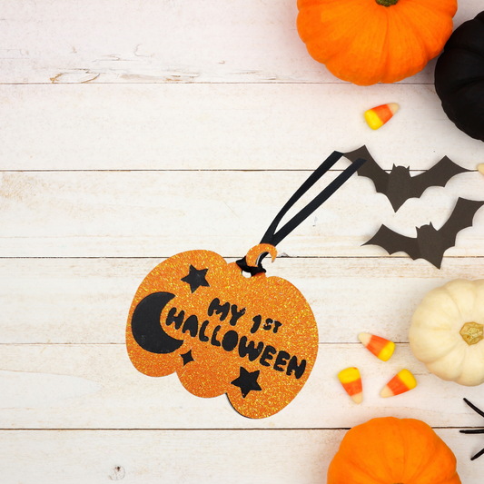 A glittery orange pumpkin-shaped Halloween Gift Tag with lettering that reads 'MY 1ST HALLOWEEN' and black hanging ribbon