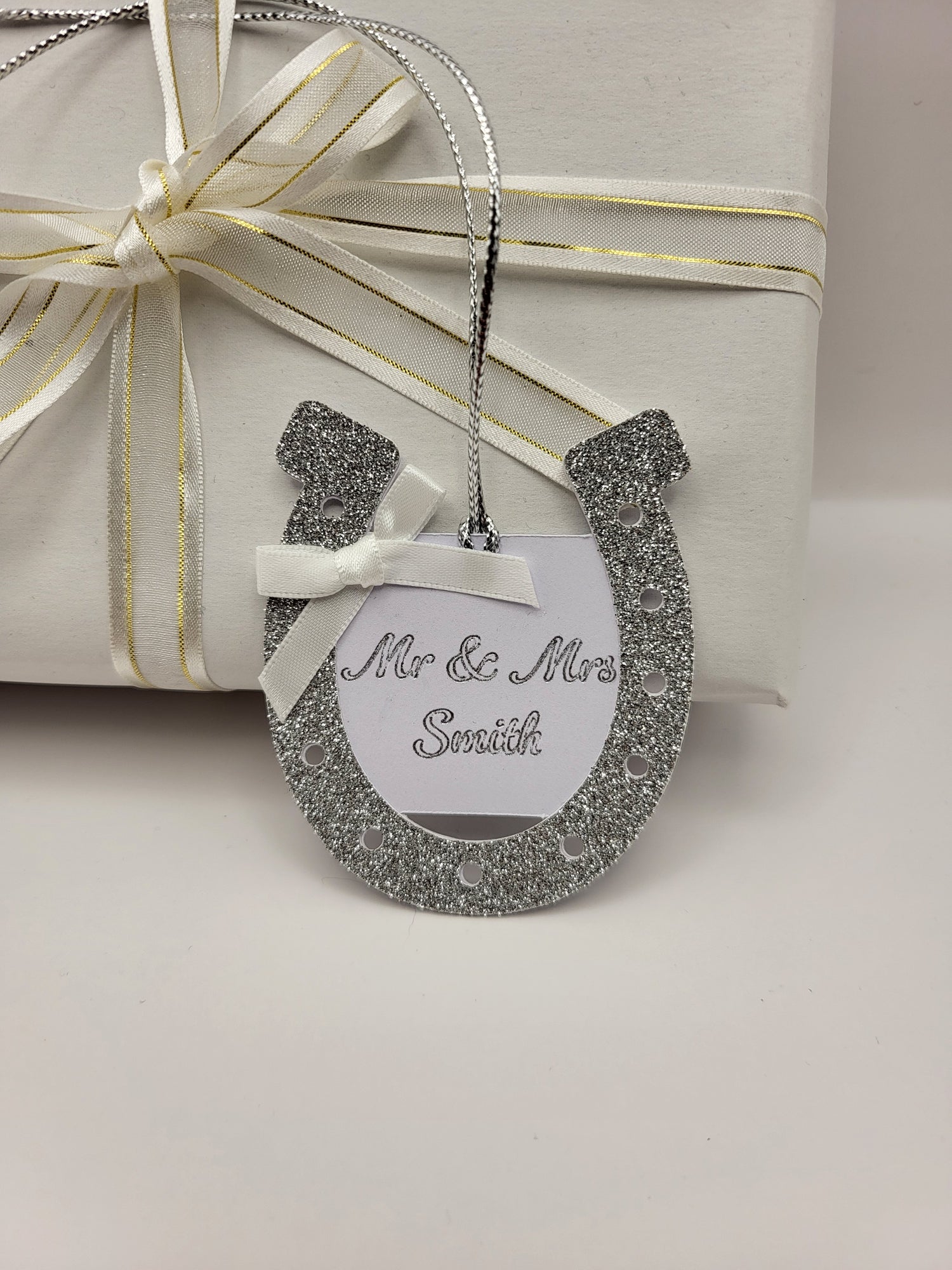 silver glitter card horseshoe shape gift tag. There is a white card centre with the words Mr &  Mrs Smith. This can be personalised into any surname you require. There is a white bow at the top left had side of the horseshoe. There is a silver metallic cord attached so you can attach it to your gift