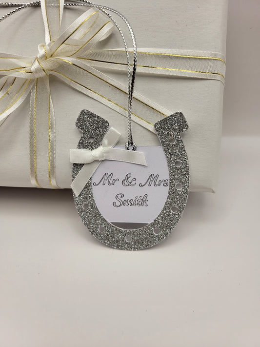 silver glitter card horseshoe shape gift tag. There is a white card centre with the words Mr &  Mrs Smith. This can be personalised into any surname you require. There is a white bow at the top left had side of the horseshoe. There is a silver metallic cord attached so you can attach it to your gift