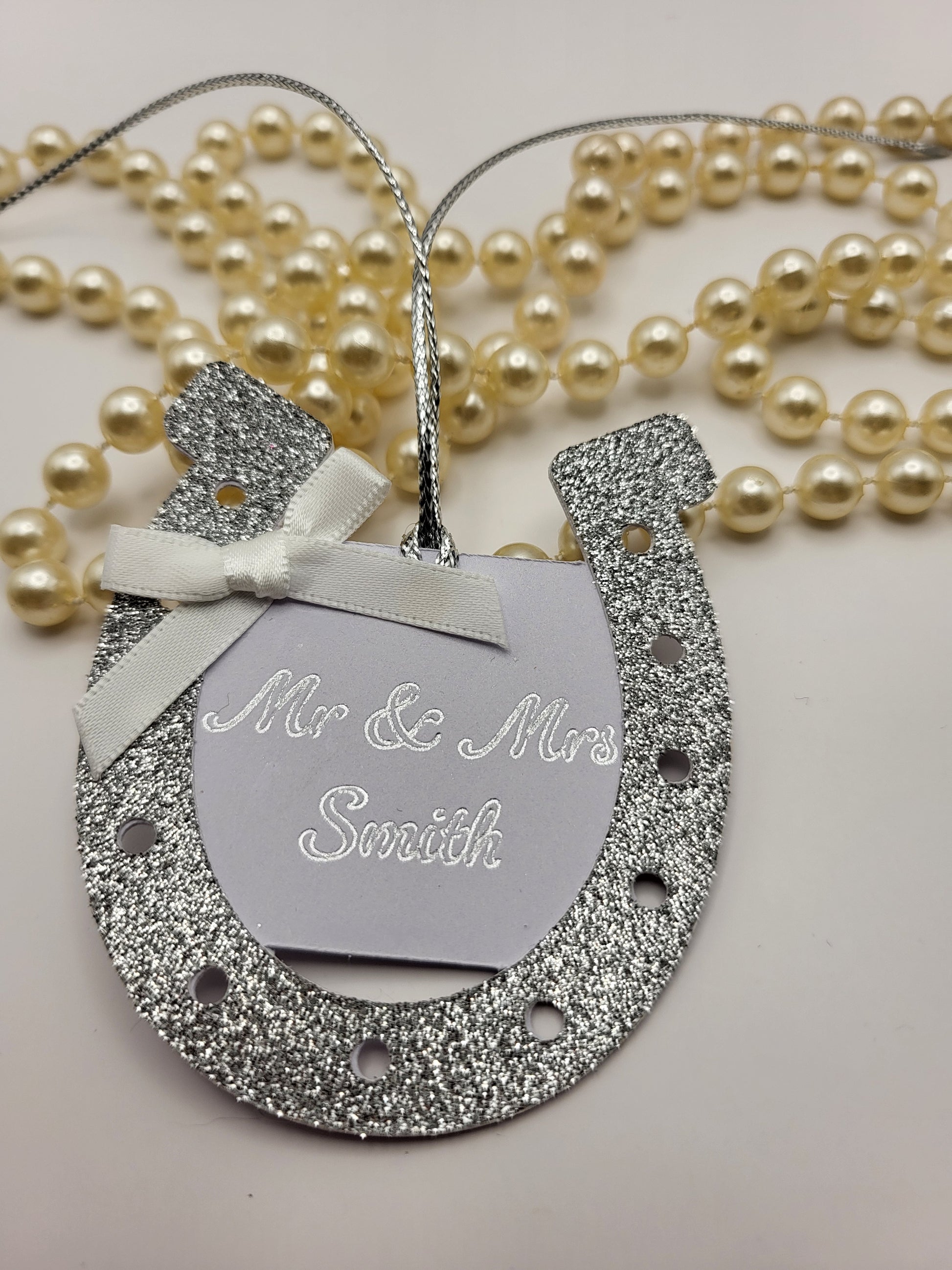 silver glitter card horseshoe shape gift tag. There is a white card centre with the words Mr &  Mrs Smith. This can be personalised into any surname you require. There is a white bow at the top left had side of the horseshoe. There is a silver metallic cord attached so you can attach it to your gift
